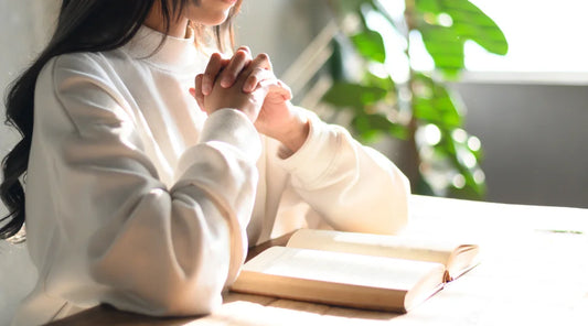 A woman sits at a table with her hands folded, embodying faith and style in a serene moment of reflection.